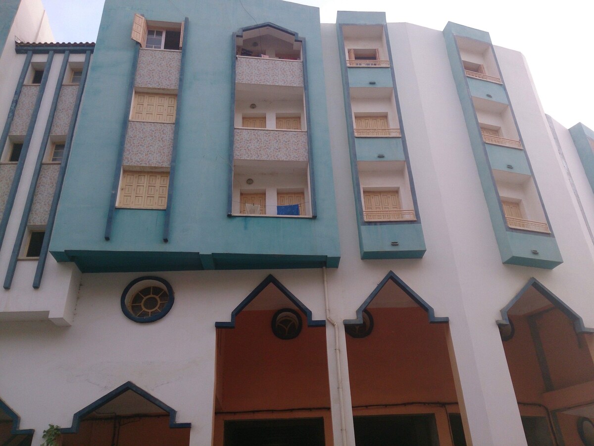 The exterior of a multi-story building features a unique architectural design with turquoise and white facades. Balconies adorned with patterned panels are visible, and the entrance showcases an arched doorway, highlighting a blend of modern and traditional elements.
