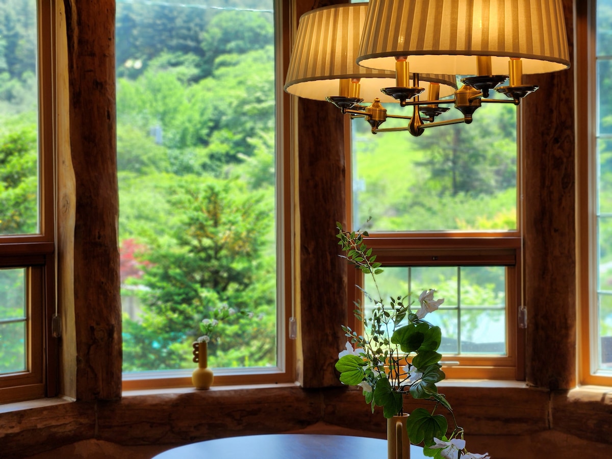Large windows allow natural light to fill the space, showcasing a stunning view of the surrounding greenery. A wooden table is adorned with a vase of fresh flowers, complemented by the warm glow of pendant lighting above.