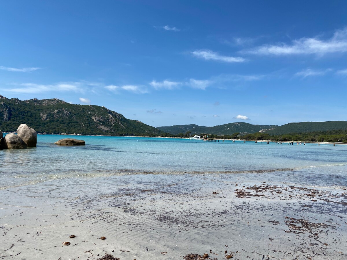 A tranquil beach scene reveals clear blue waters gently lapping against a sandy shore. Rocky formations can be seen on the left, and the landscape includes lush hills in the background, with a few distant figures enjoying the calm environment.