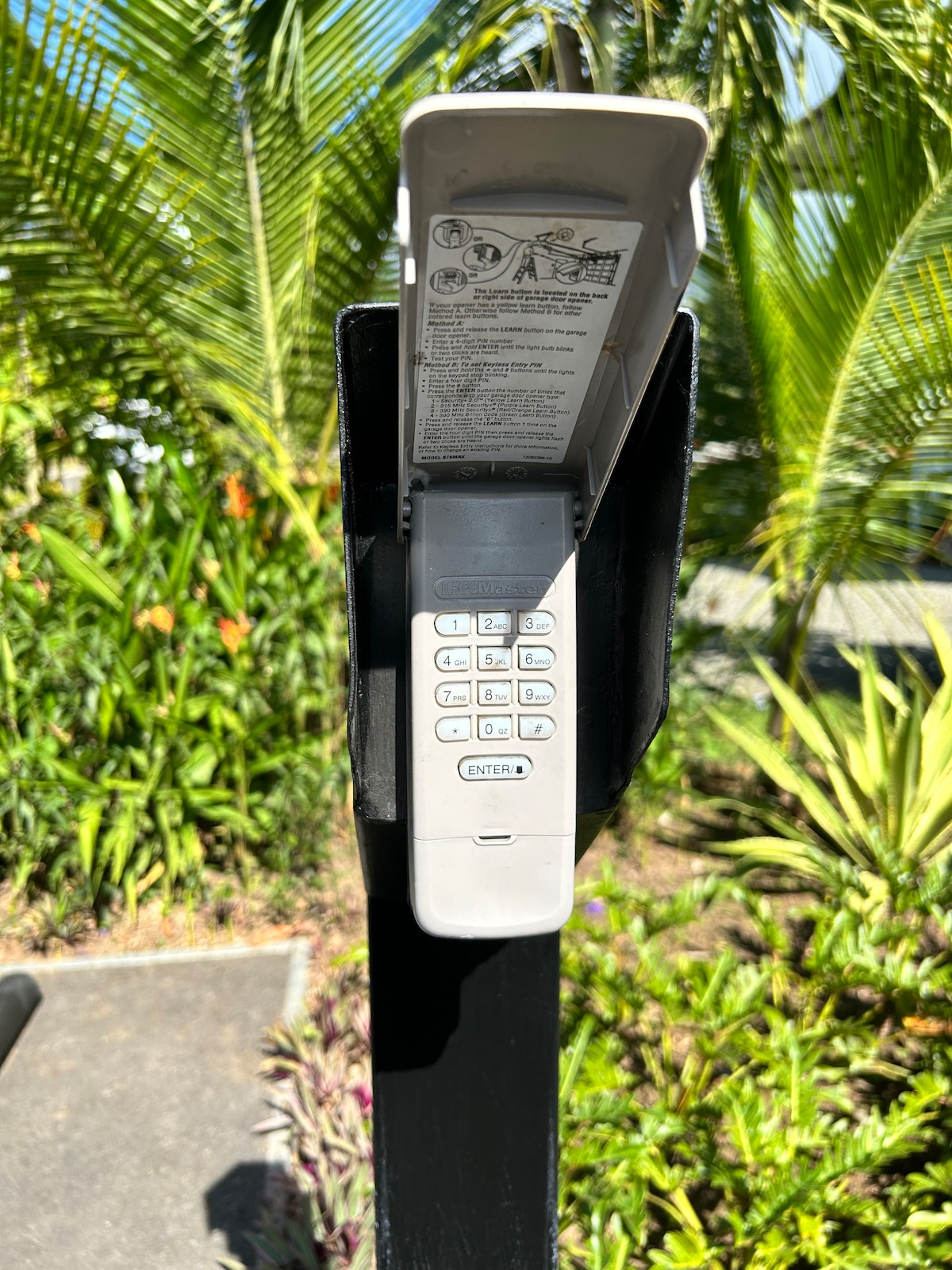 A wall-mounted intercom system is displayed, featuring a numeric keypad with clear labeling. The device is positioned against a backdrop of lush tropical greenery, including various plants and palm leaves.