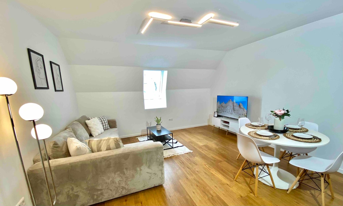 The living area features a modern sofa paired with a sleek coffee table, illuminated by stylish lighting. A flat-screen television is positioned for easy viewing, while a dining space with four chairs is arranged beside a window, allowing natural light to fill the room.