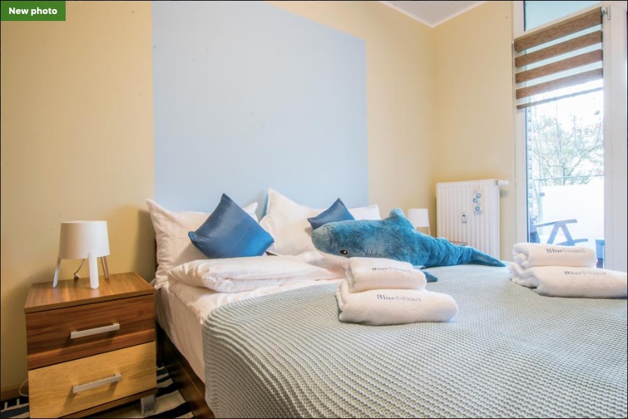 A cozy bedroom features a large bed adorned with soft linens and decorative pillows. A plush shark-shaped toy rests on the bed. Flanking the bed are two bedside tables, each with a lamp. Natural light flows in through a window, providing a serene ambiance.