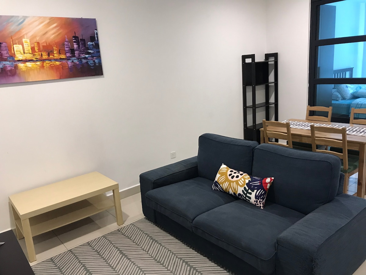A living area features a dark blue sectional sofa with a decorative cushion, accompanied by a light wood coffee table. A painting of a city skyline decorates the wall above the sofa. A dining area with a wooden table and chairs is visible in the background.