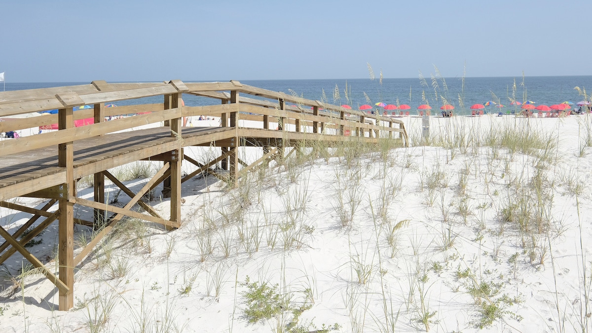 A wooden boardwalk leads through sandy dunes, surrounded by patches of grass. The boardwalk provides access to the beach, where colorful umbrellas are set up in the distance, contrasting against the blue ocean and clear sky.