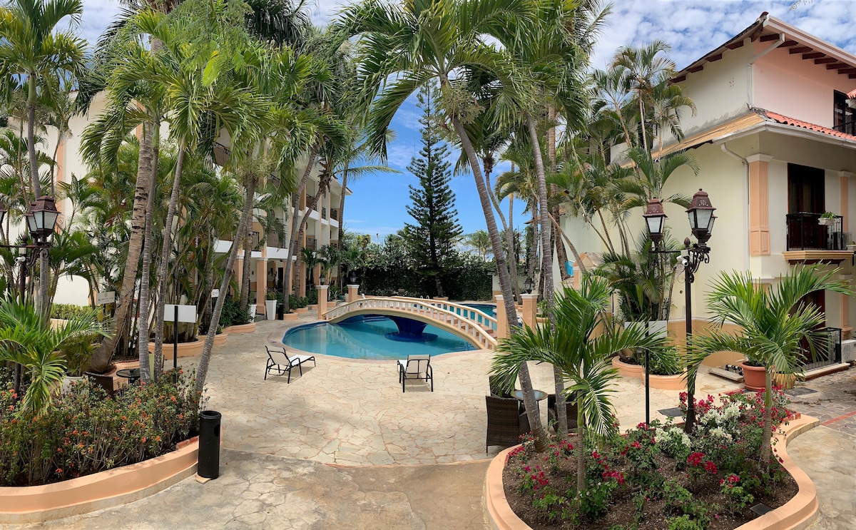 A serene pool area is surrounded by palm trees and lush greenery. Two lounge chairs are positioned by the water, with a patio offering ample space for relaxation. The surrounding buildings reflect a charming architectural style, contributing to a tranquil atmosphere.