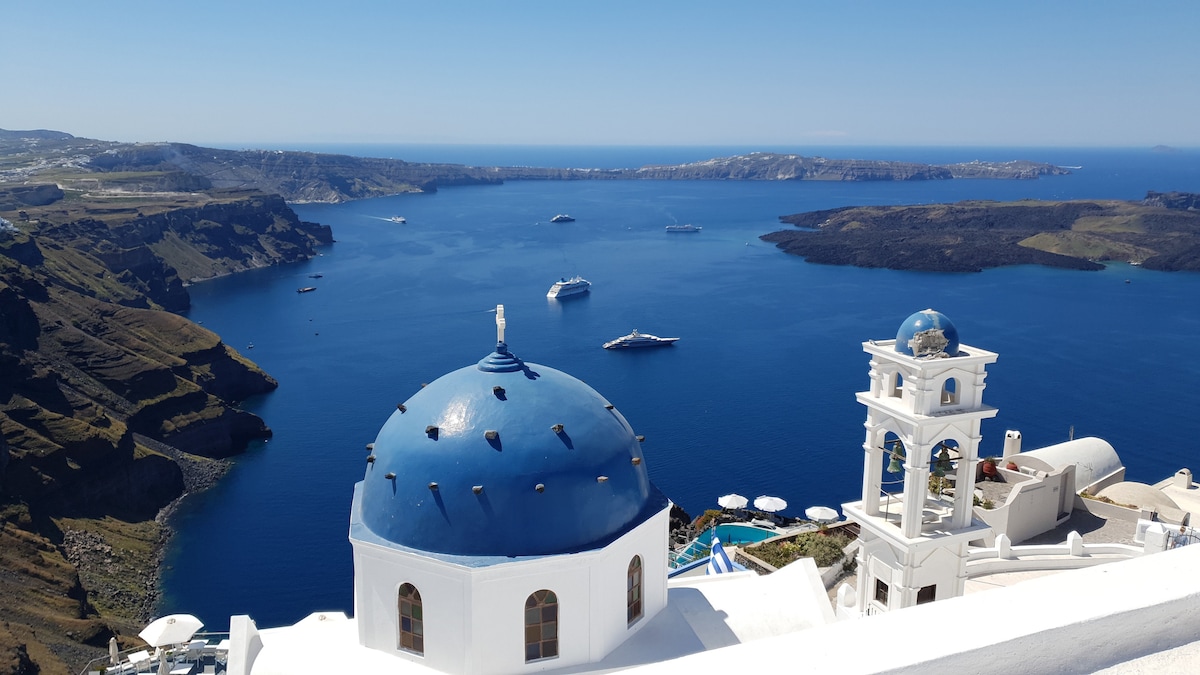 The image showcases a stunning view over the caldera, with a vibrant blue dome of 'Anastasi church' in the foreground. The deep blue water is dotted with boats, while the rugged cliffs of Santorini rise dramatically in the background under a clear sky.