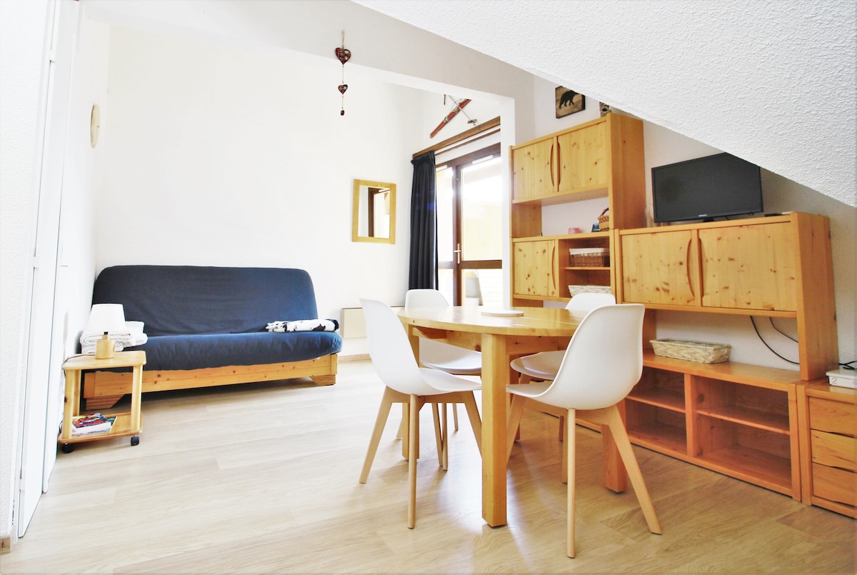 A bright living space features a convertible sofa bed and a round wooden dining table surrounded by four white chairs. Light wood furniture lines the room, while a large window provides natural light and opens to a balcony. A flat-screen TV is positioned on a wooden unit.