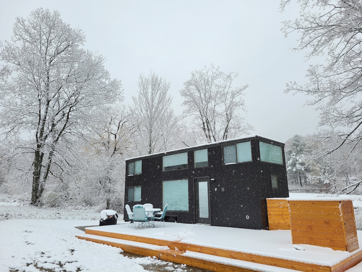 The tiny house is surrounded by a snowy landscape, with snow-covered trees in the background. A small deck is visible, featuring a round table and chairs. The black exterior contrasts with the white snow, creating a peaceful winter scene.