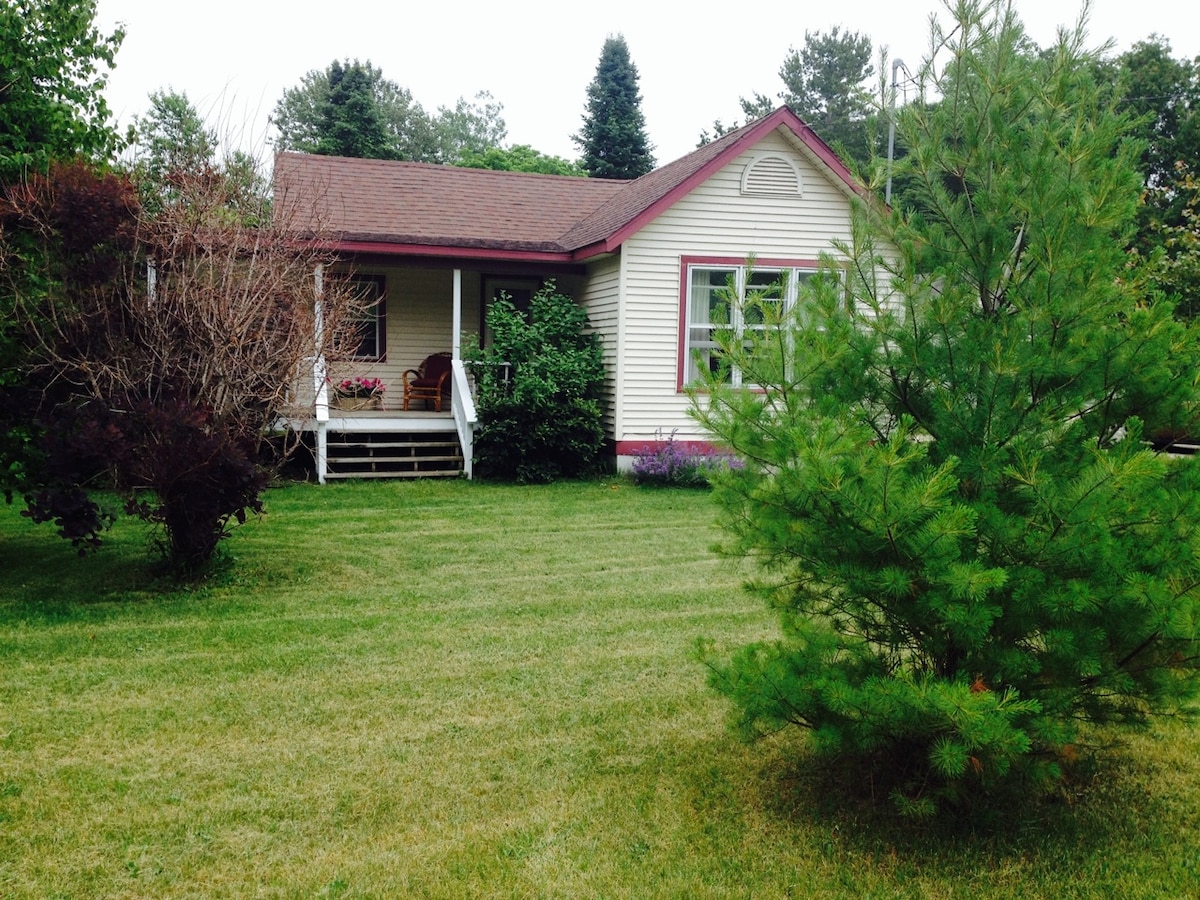 A well-maintained cottage is surrounded by lush green grass and trees, featuring a welcoming front porch with seating. The exterior showcases light-colored siding with dark trim, complemented by vibrant landscaping, creating a cozy and inviting appearance.