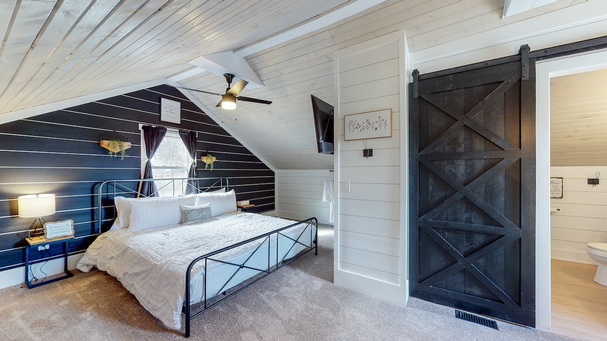 An inviting master suite is showcased, featuring a spacious bed with soft bedding against a backdrop of dark accent walls. A ceiling fan provides airflow, while natural light floods the room. A sliding barn door leads to a modern bathroom, adding a touch of rustic elegance.