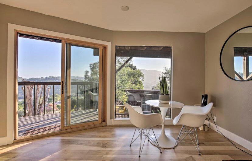 A dining area is highlighted by a round table surrounded by white chairs. Large glass doors open to the deck, providing views of the surrounding landscape. A circular mirror reflects the soft interior light, complementing the light-colored walls and wooden flooring.
