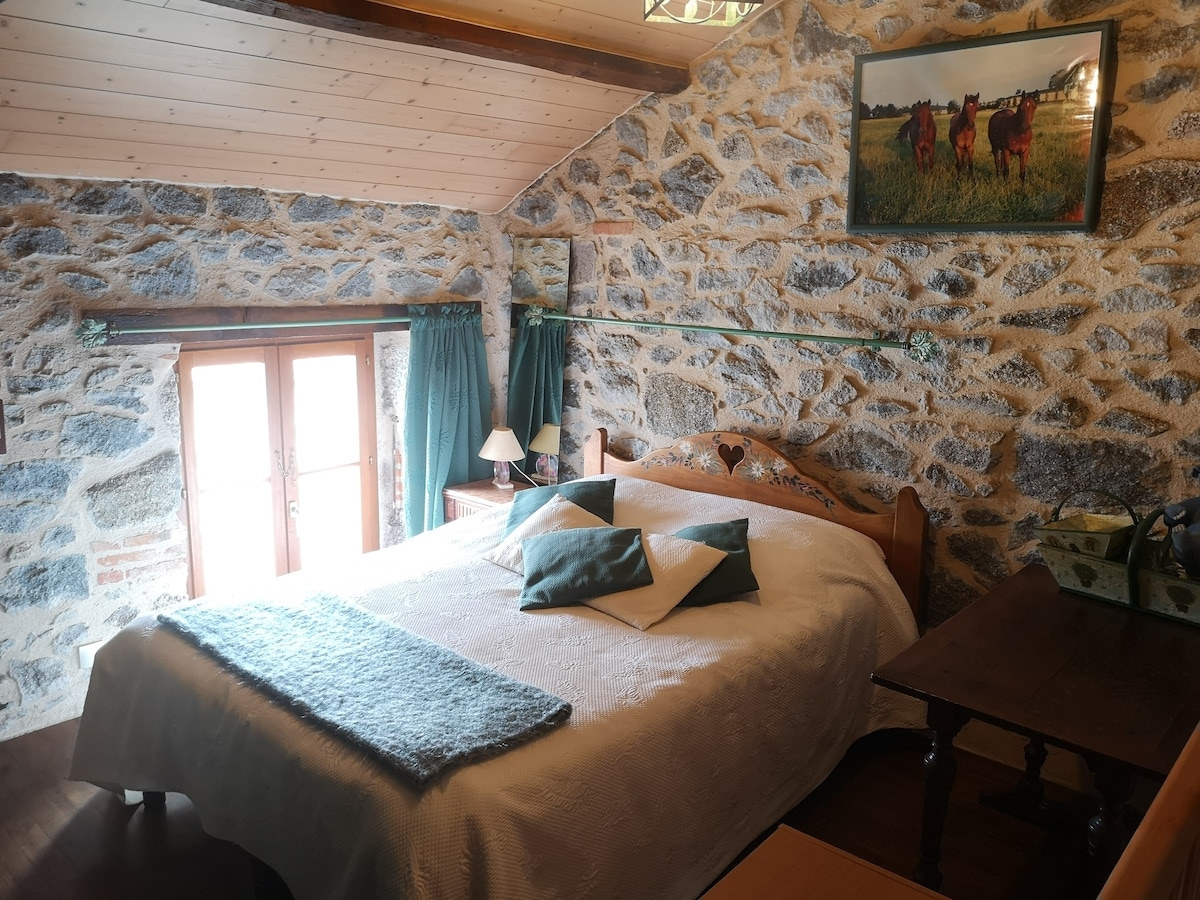A cozy bedroom features a spacious bed dressed in white linens, adorned with decorative pillows. Stone walls provide texture, while natural light enters through two windows with green curtains. A wooden table is positioned nearby, complemented by soft lighting from bedside lamps.