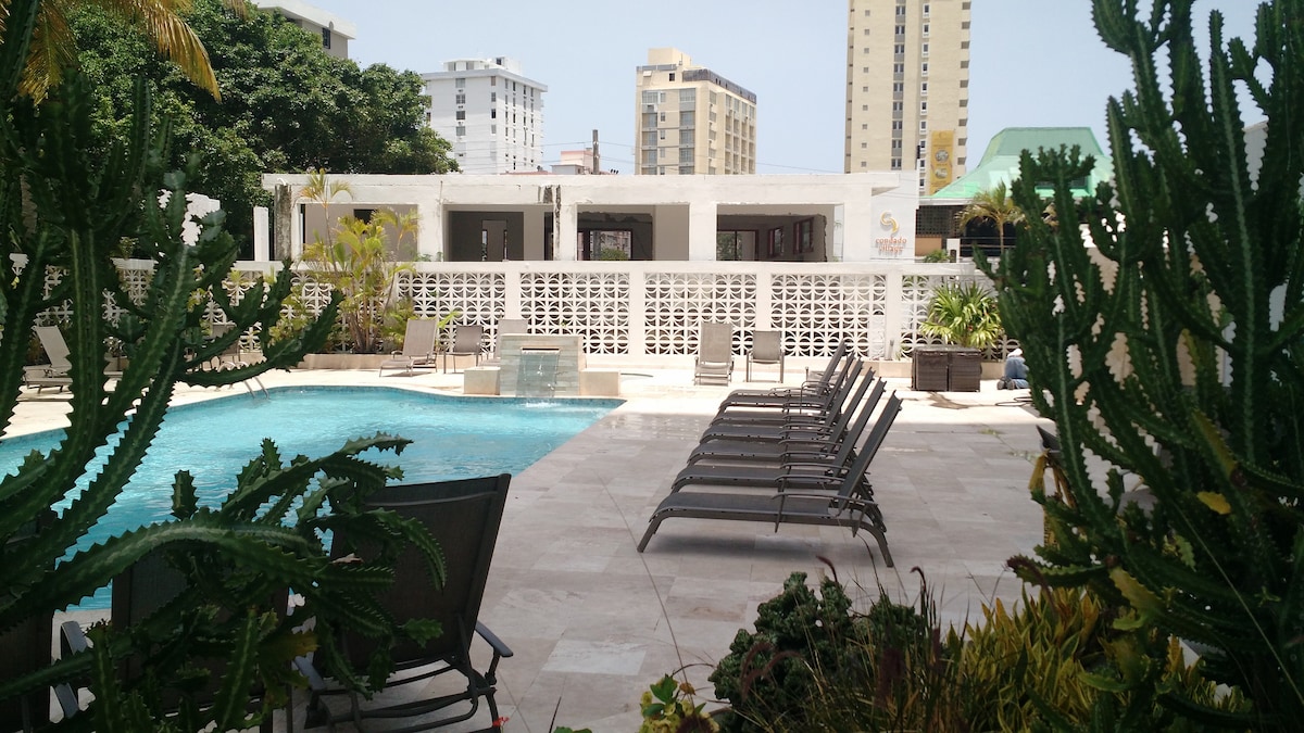 A well-maintained swimming pool area is framed by lush greenery and modern lounge chairs. The pool's serene water surface reflects surrounding buildings, with minimalistic decor enhancing the outdoor space. Sunlight creates a bright and open atmosphere, inviting relaxation.