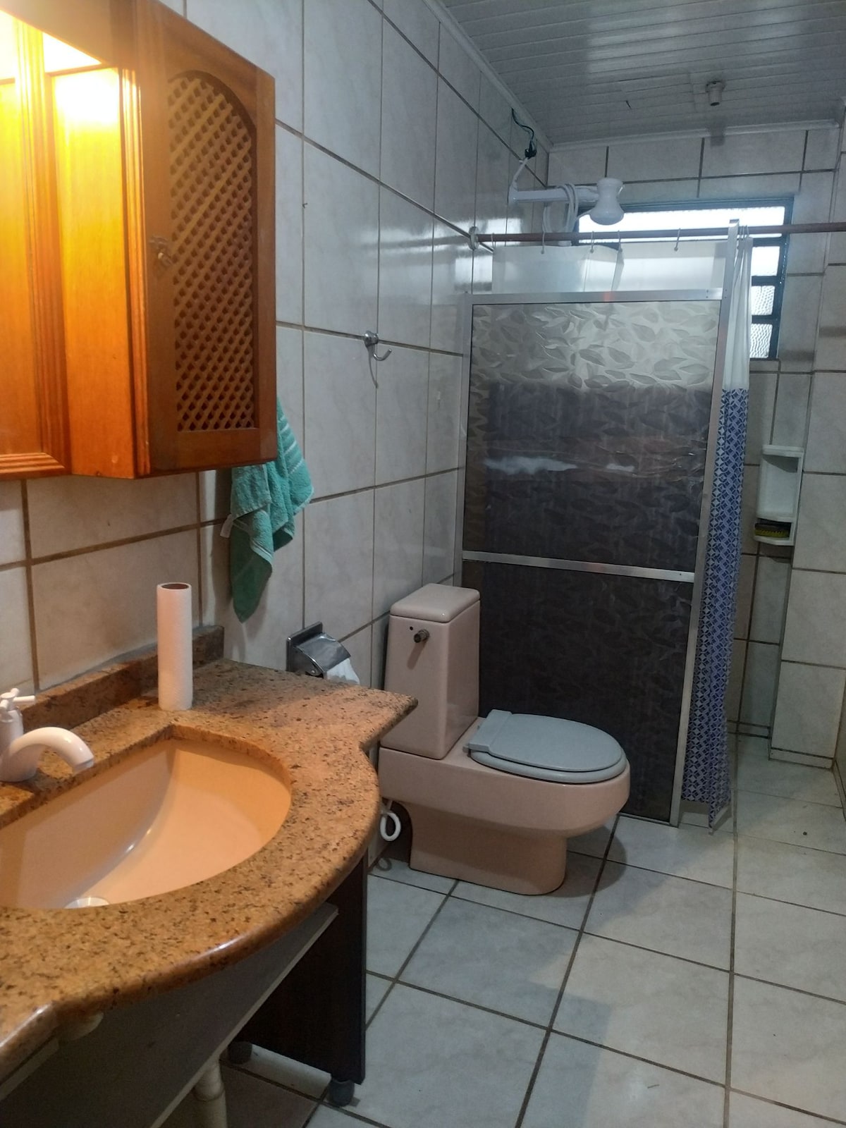 A clean bathroom features tiled walls and a spacious shower area, enclosed by a glass partition. A granite countertop holds a sink, complemented by a wooden cabinet above. A towel is hung on a hook nearby, and a roll of toilet paper is placed within easy reach.