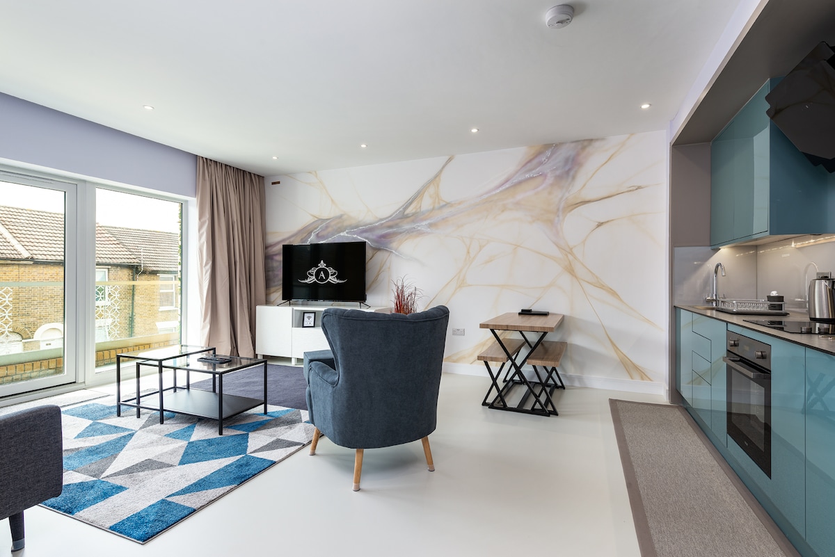 A modern living space features a large window allowing natural light to fill the room. A grey armchair and black coffee table are positioned on a stylish blue and grey rug. A wall-mounted television is visible along with a decorative wall mural, enhancing the contemporary design.