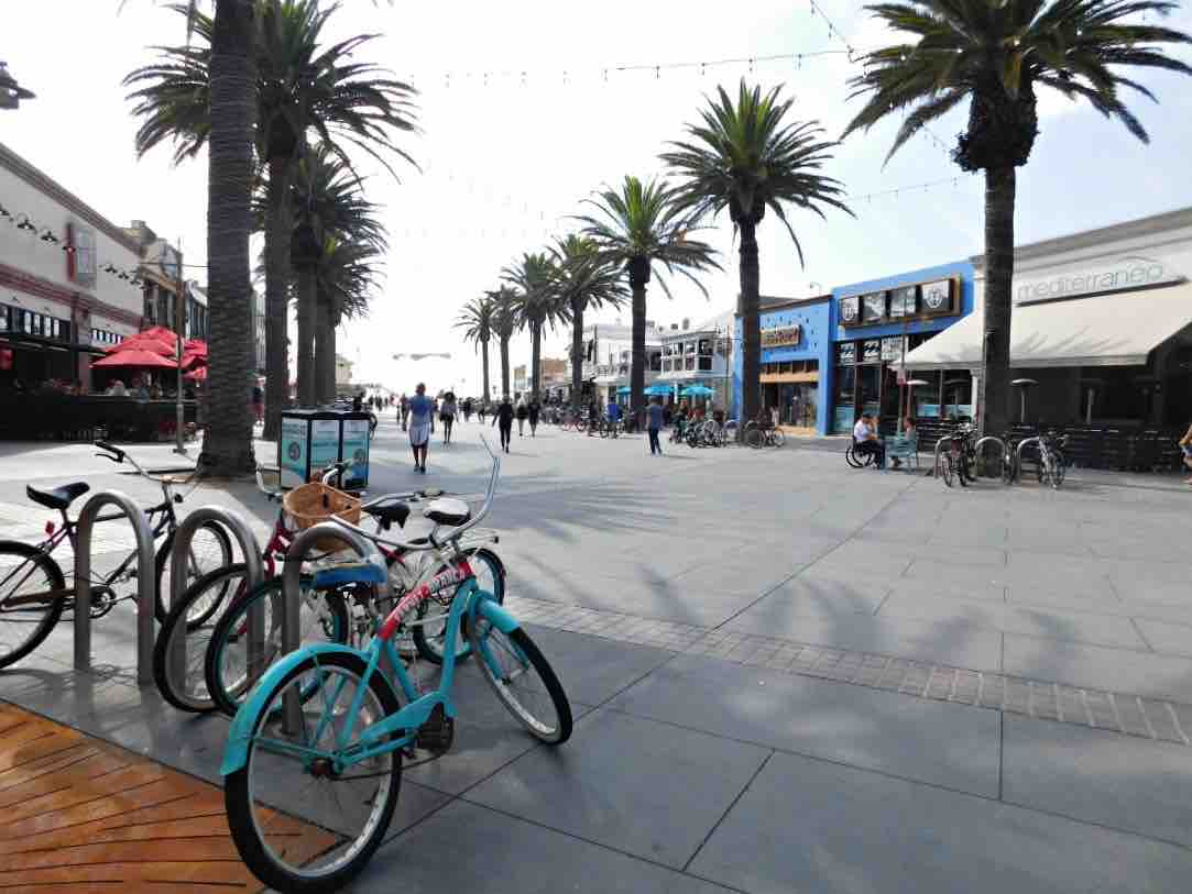 A lively street scene features a wide walkway lined with palm trees on either side. Bicycles are parked along the side, while people stroll in the distance, showcasing a vibrant atmosphere near shops and dining establishments.