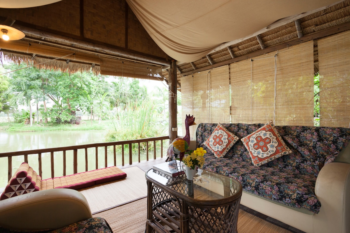 A cozy sitting area features a floral-patterned sofa with colorful cushions, complemented by a wicker coffee table. Natural light filters through bamboo blinds, creating a serene ambiance. The view showcases a lush garden and a tranquil pond, enhancing the peaceful outdoor connection.