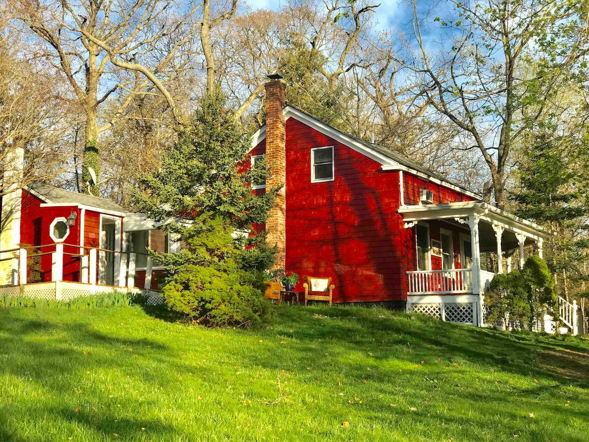 A charming red cottage is set among lush greenery, showcasing a manicured lawn leading to the porch. The house features a rustic stone chimney and a welcoming front porch adorned with white railings. Nearby, a small structure complements the main home with a rounded window.