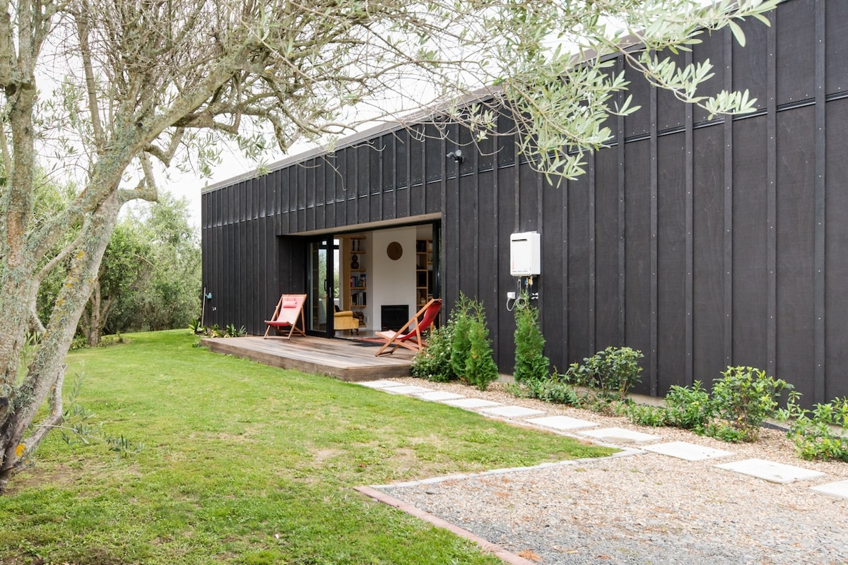 A modern black-clad home is surrounded by lush greenery, with a wooden deck featuring two red lounge chairs. A stone path leads from the driveway, which is bordered by trimmed hedges and garden beds, enhancing the outdoor living space.