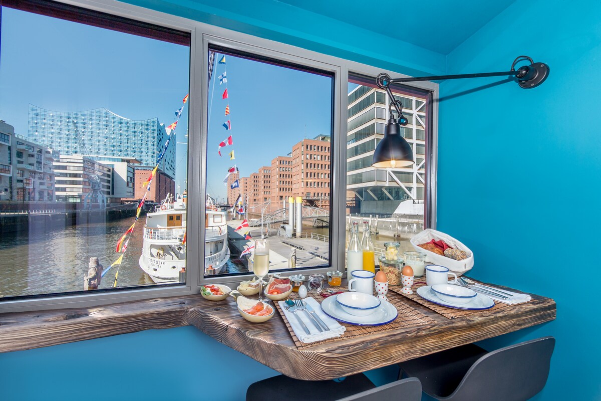 A dining area is positioned beside large windows, providing a view of the vibrant harbor. The table is set with breakfast items, including pastries, fruit, and drinks. The bright blue walls and natural wood table create a welcoming atmosphere, complemented by modern light fixtures.