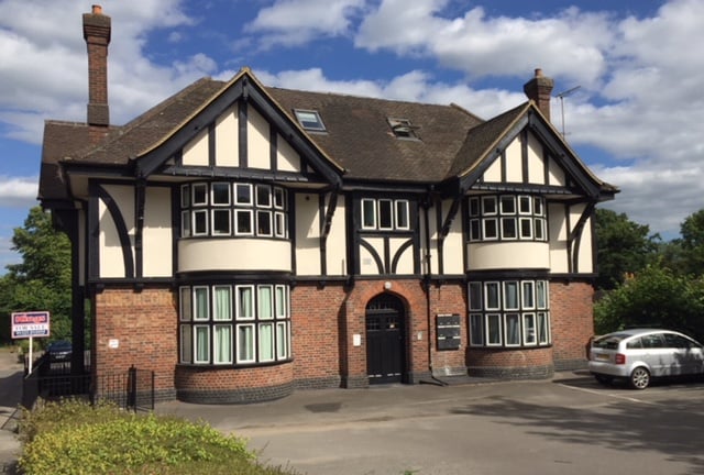 A two-story building featuring a distinctive Tudor-style design with white and black accents. Large, paned windows dominate the facade, allowing plenty of natural light. The entrance is framed by a decorative archway, with a gravel area visible in front, providing space for parking.