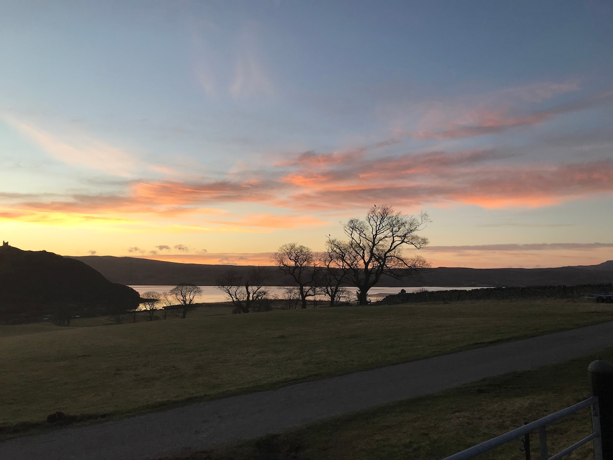 A serene landscape is captured at sunset, featuring a calm body of water reflecting soft pink and orange hues in the sky. Silhouettes of trees are visible against the distant hills, creating a tranquil atmosphere. A well-kept grassy area leads towards the water's edge.