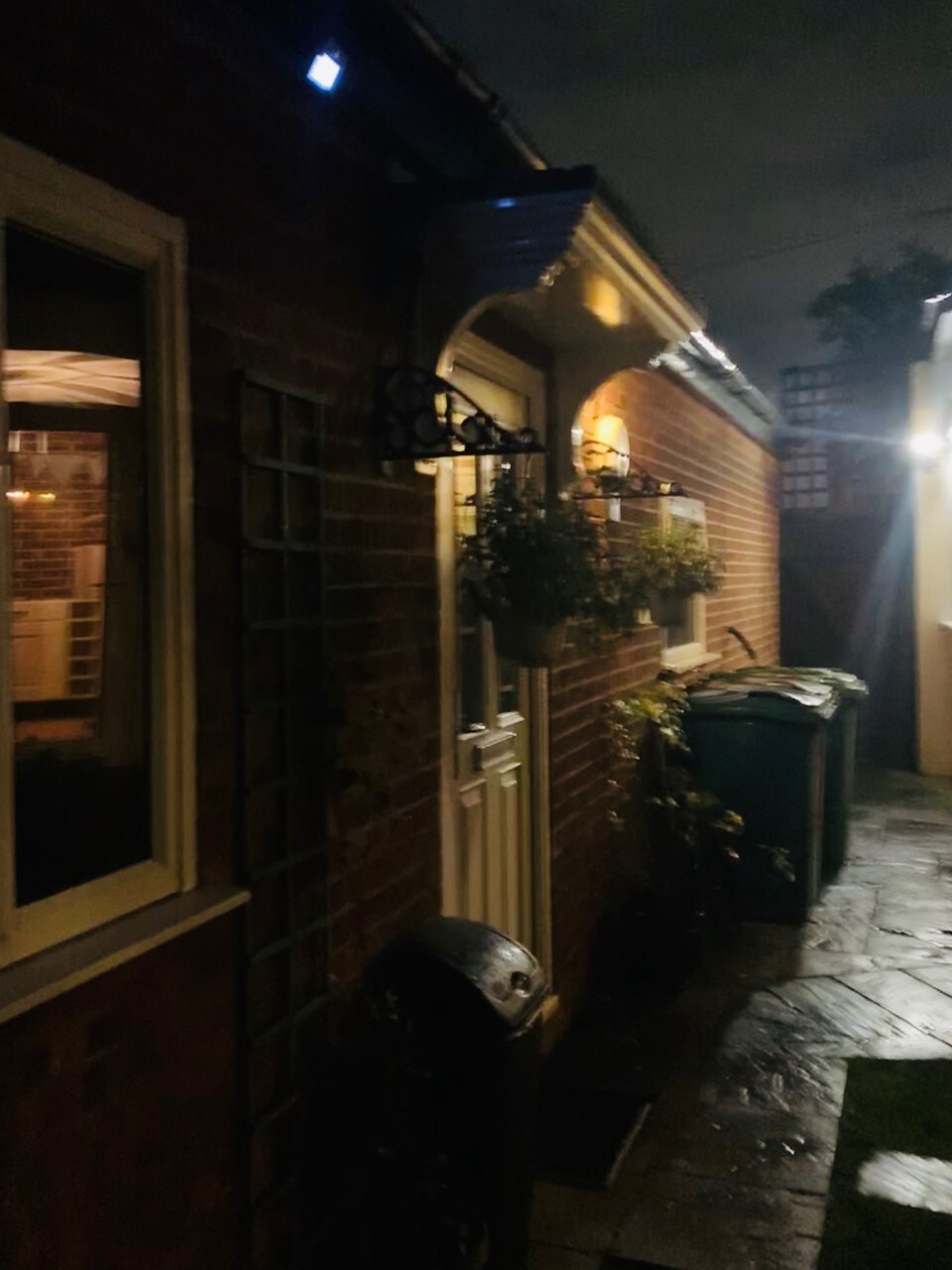 An exterior view of the studio is displayed at night, with soft lighting highlighting the brick facade. A white door with simple trim is framed by a small decorative element. Nearby, a green bin and stone pathway are visible, along with a light illuminating a corner.