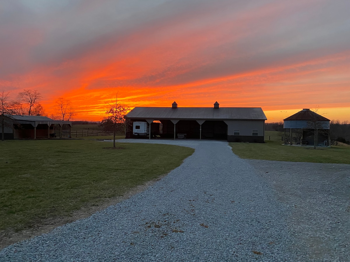 A vibrant sunset casts shades of orange and pink across the sky, illuminating the spacious property. A gravel driveway leads up to a barn structure, flanked by green grass and a small gazebo. Silhouettes of trees are visible in the background.