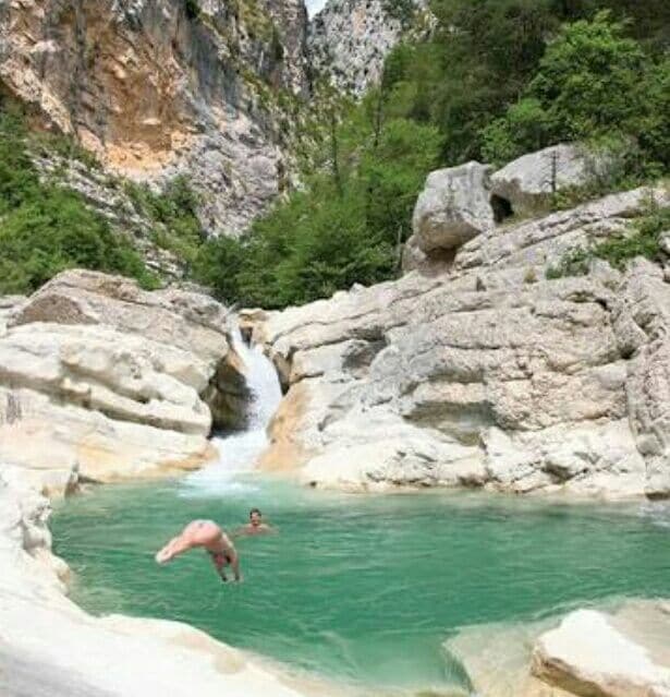 A natural swimming area is shown, featuring clear turquoise water surrounded by smooth, light-colored rock formations. A person is diving into the water, while another person swims nearby. Lush greenery and rocky cliffs provide a scenic backdrop, emphasizing the tranquil setting.