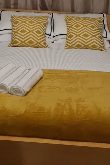 The inviting sleeping area features a spacious bed adorned with a textured light-colored blanket. Two decorative pillows with geometric patterns provide a pop of color, complemented by neatly arranged rolled towels at the foot of the bed.