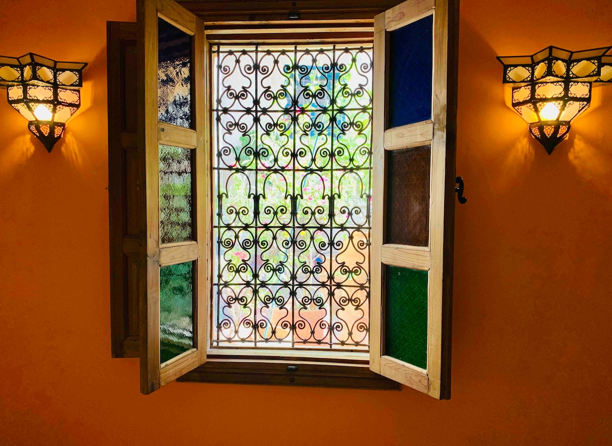 A vibrant window is framed by wooden shutters, showcasing ornate ironwork. Colorful glass panels allow glimpses of the lush greenery outside. Two decorative wall sconces illuminate the warm orange walls, enhancing the cozy ambiance of the space.