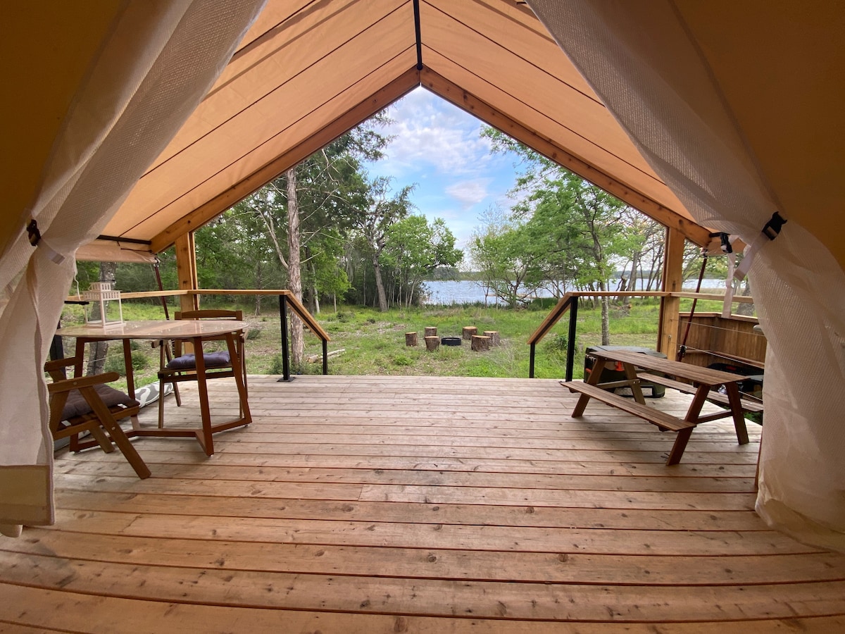 The entrance of the safari-style tent opens to a wooden deck with seating arrangements. Nearby, a firepit area is visible against a backdrop of lush greenery and the lake, inviting outdoor gatherings and relaxation.