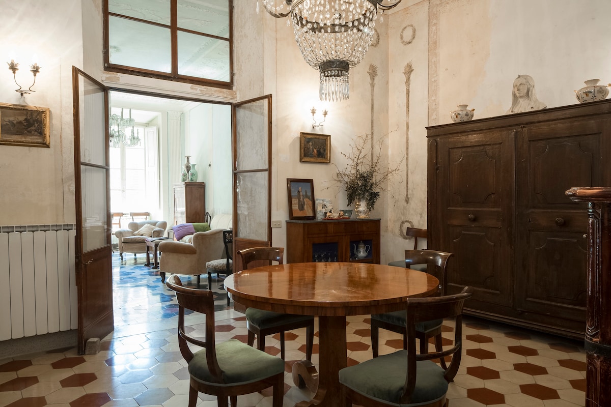 A dining area features a round wooden table surrounded by four wooden chairs, set against a backdrop of vintage decor. An antique cabinet and elegant chandelier add character, while light enters through oversized doors leading to a spacious seating area visible in the background.