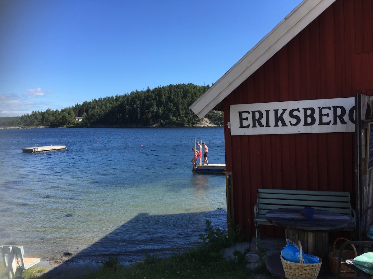 A red building labeled 'ERIKSBERG' is featured beside a calm blue water area. People are seen on a wooden dock, enjoying the water. The background shows lush greenery and distant shoreline, conveying a tranquil natural setting.