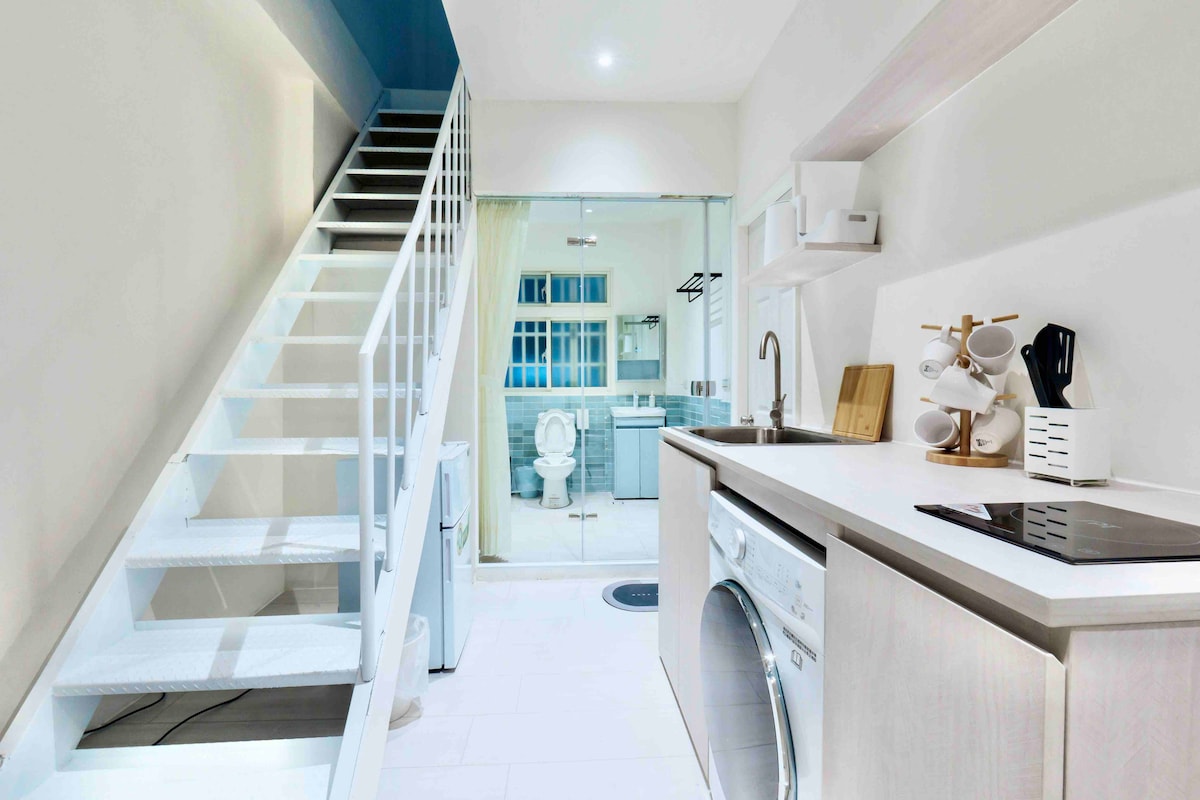 A modern kitchenette features a compact layout with a stovetop and washing machine. Open shelving displays utensils, while a staircase leads to the upper level. A glass partition provides a view of the bathroom area, contributing to an open and functional design.