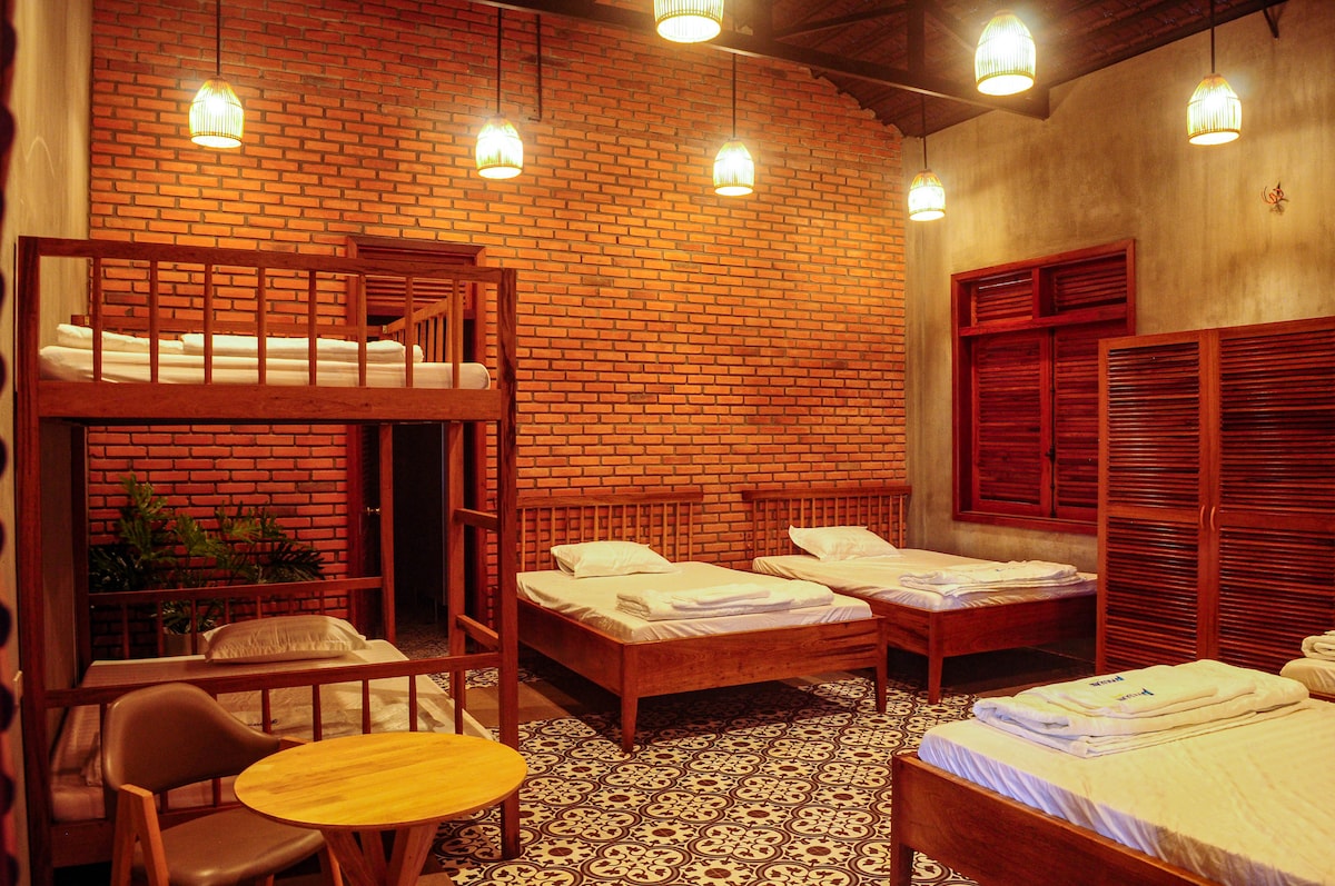 A spacious room features multiple bunk beds and single beds arranged for group accommodation. Wooden furniture, including a small round table and chairs, complements the warm brick walls. Soft lighting from hanging fixtures creates a cozy atmosphere, while patterned tile flooring adds visual interest.