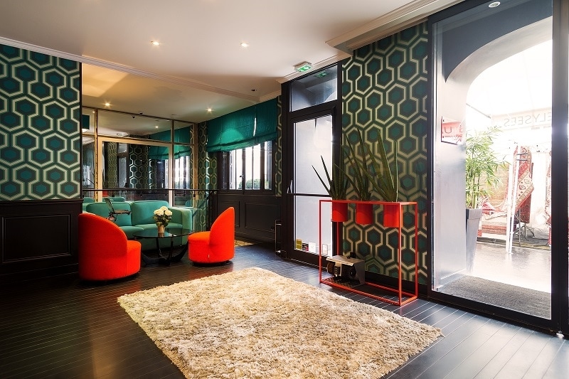 A modern lobby area features bold geometric wallpaper in green and gold tones. Two bright red chairs provide seating around a light beige rug. Large windows allow natural light to fill the space, while decorative plants add a touch of greenery.