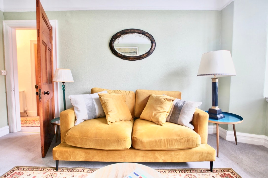 A welcoming living area is highlighted by a plush yellow sofa adorned with soft cushions. A round side table complements the space, while a vintage mirror hangs above, reflecting the natural light from the nearby window. A door leads to another room in the background.
