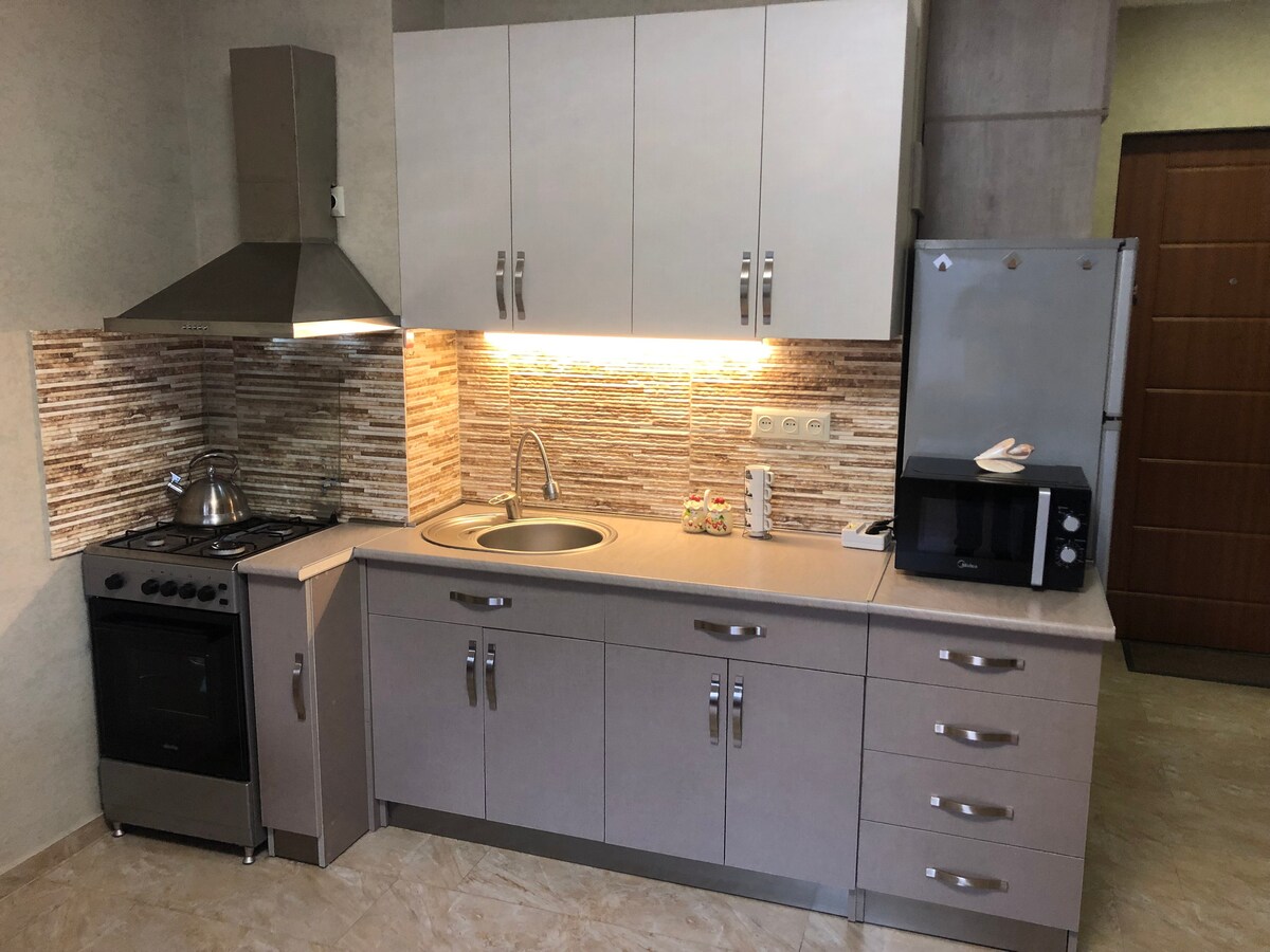 A modern kitchen space features sleek cabinetry and a stone accent wall. Appliances include a stove, microwave, and refrigerator. A stainless steel sink is positioned under under-cabinet lighting, highlighting the functional layout suitable for meal preparation.