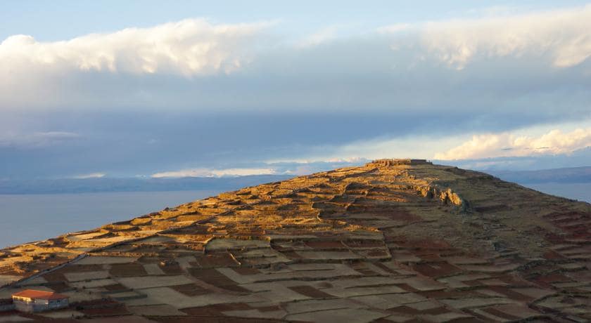 A panoramic view of a terraced hillside with various plots of cultivated land. The landscape features earthy tones of brown and beige, contrasting with a calm body of water in the distance. Soft light reflects off the clouds, creating a serene and expansive backdrop.
