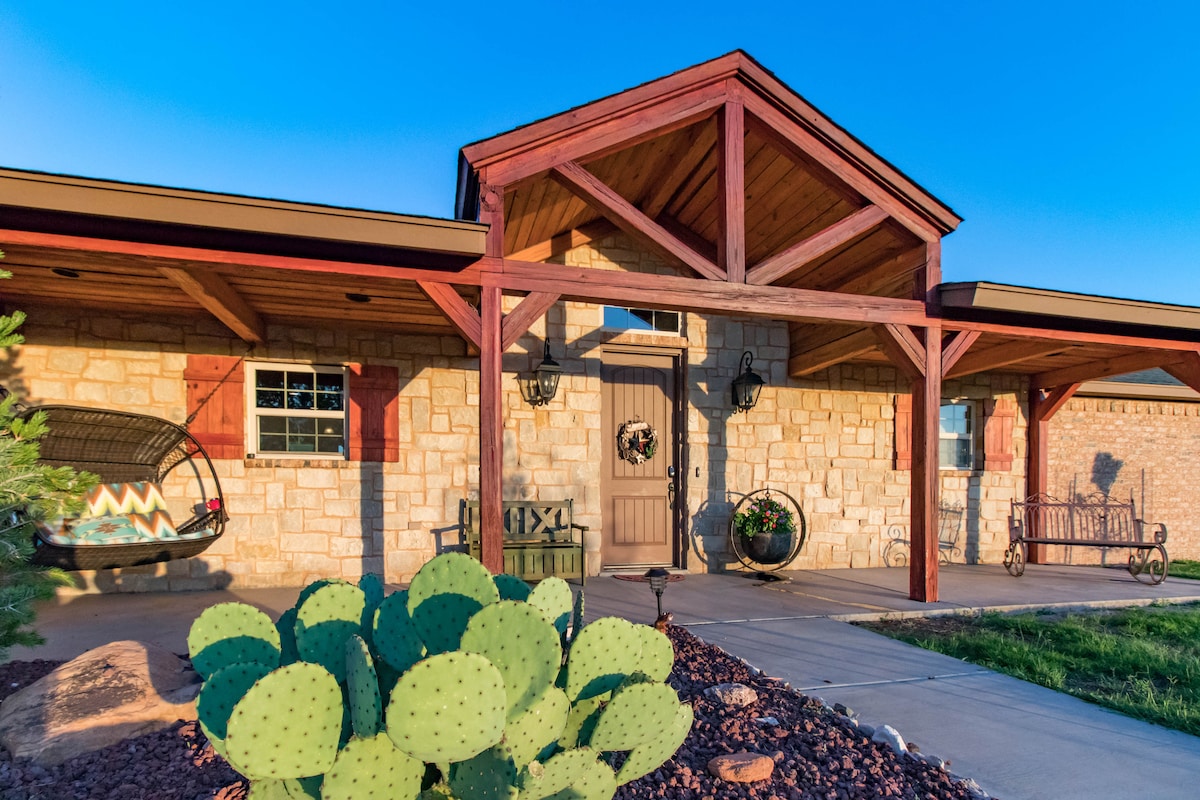 A charming lodge entrance is framed by a wooden porch supported by decorative beams. The stone facade complements the natural landscape, with a vibrant cactus positioned prominently in the foreground. A rustic swing and a bench are visible, inviting relaxation in a serene setting.