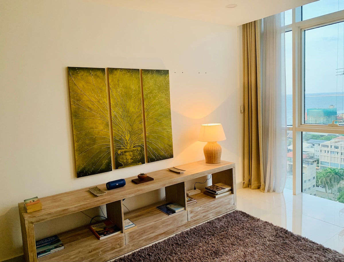 A spacious living area features a wooden console with an assortment of books displayed. A textured rug adds warmth to the room, which is illuminated by a soft lamp. Large windows offer views of the ocean and city, complemented by sheer curtains.