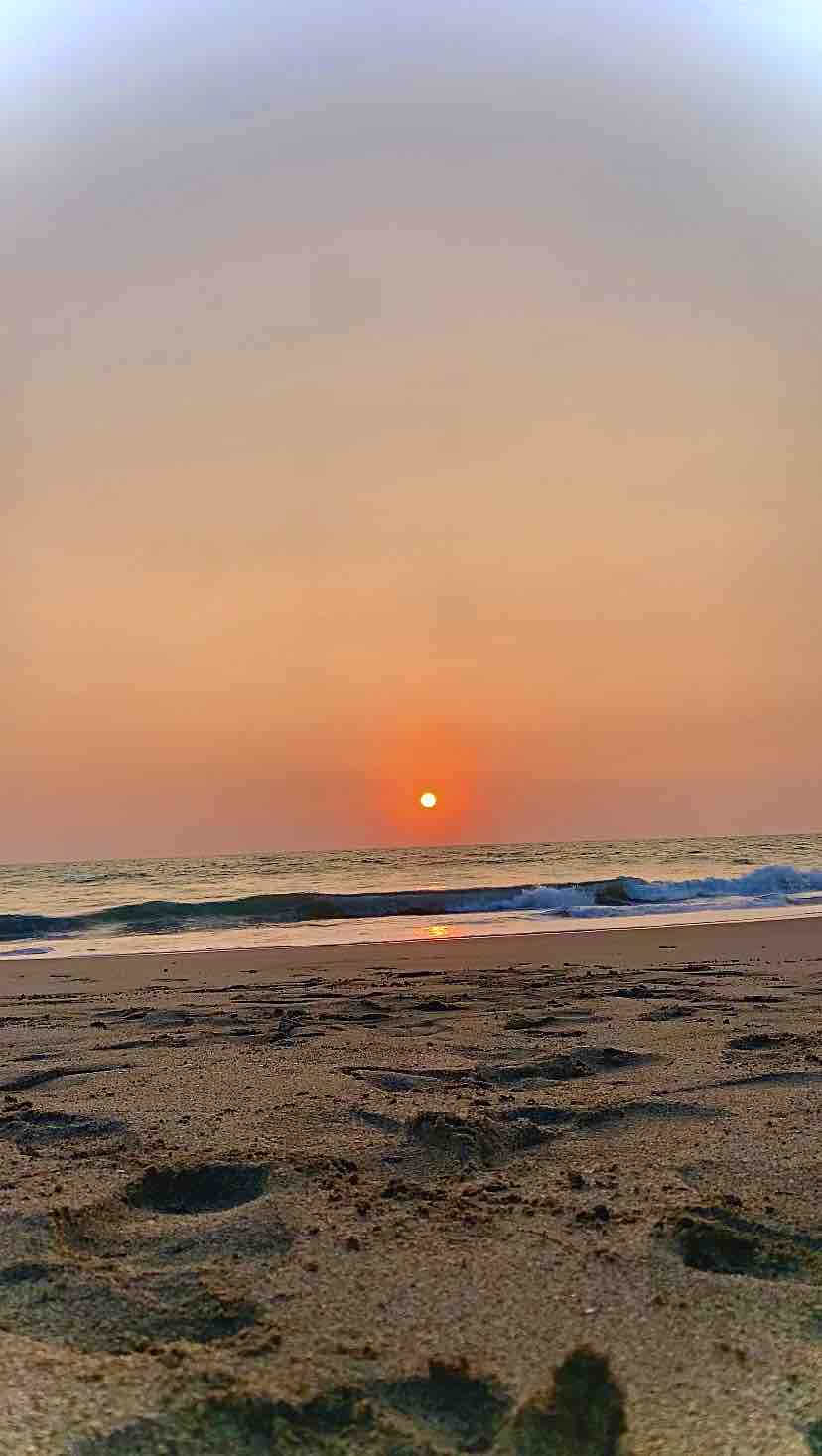 A serene beach scene captures the sun setting over the horizon, casting a warm glow on the surface of the water. Gentle waves create a rhythmic motion along the sandy shore, while footprints are visible in the foreground, leading towards the tranquil sea.