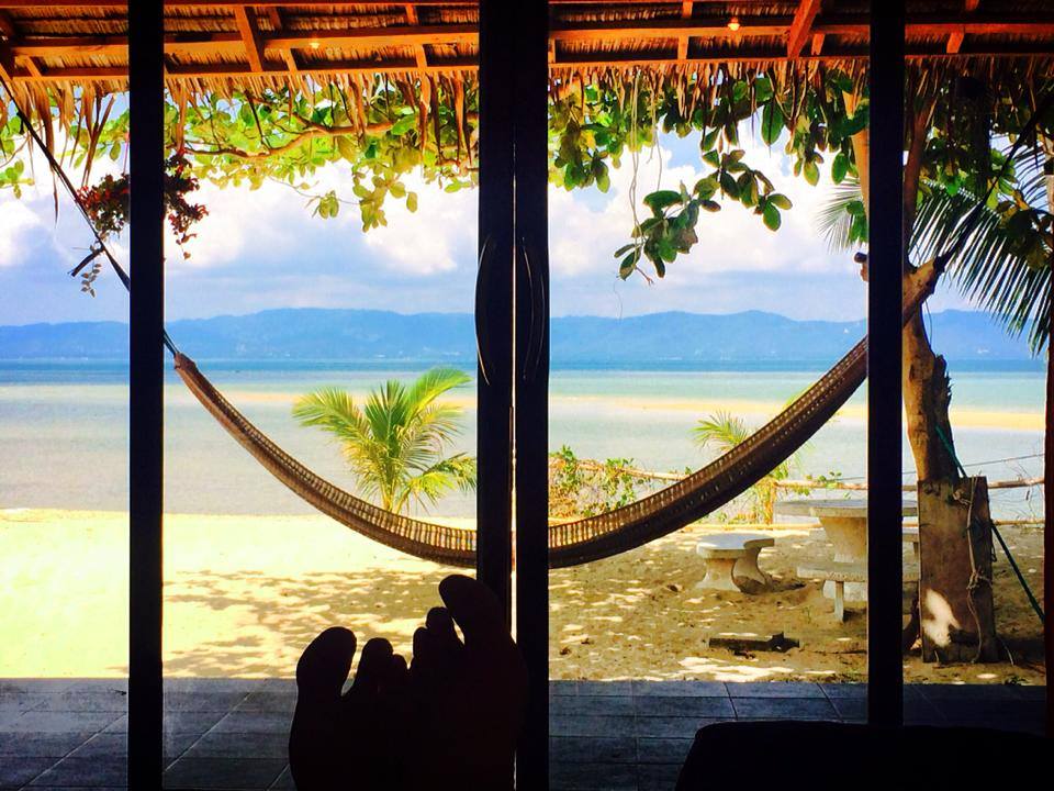 A hammock is viewed from an open doorway, framed by natural greenery. The calm water extends toward distant mountains under a partly cloudy sky. A sandy area is visible, complemented by a stone table and benches in the foreground.