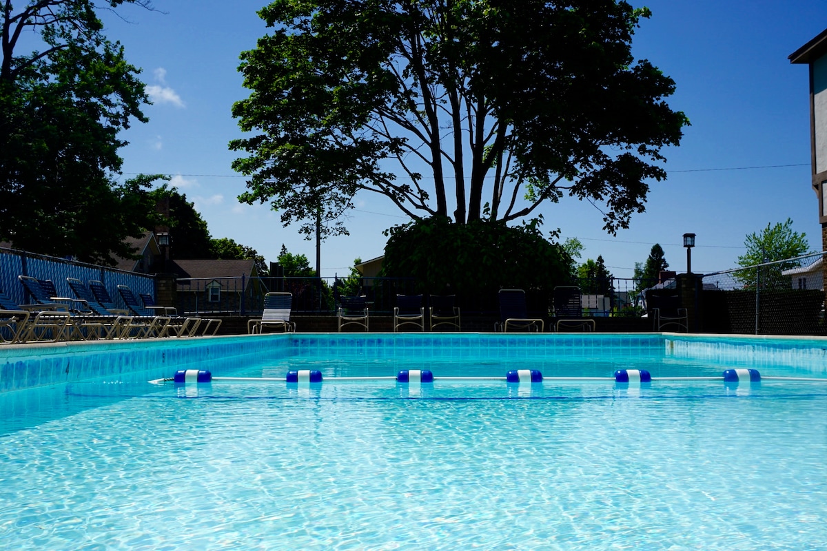 A seasonal outdoor pool is accessible, featuring clear turquoise water and marked by blue and white lane dividers. Surrounding the pool, a spacious deck is equipped with lounge chairs, and a large tree provides shade in the background.