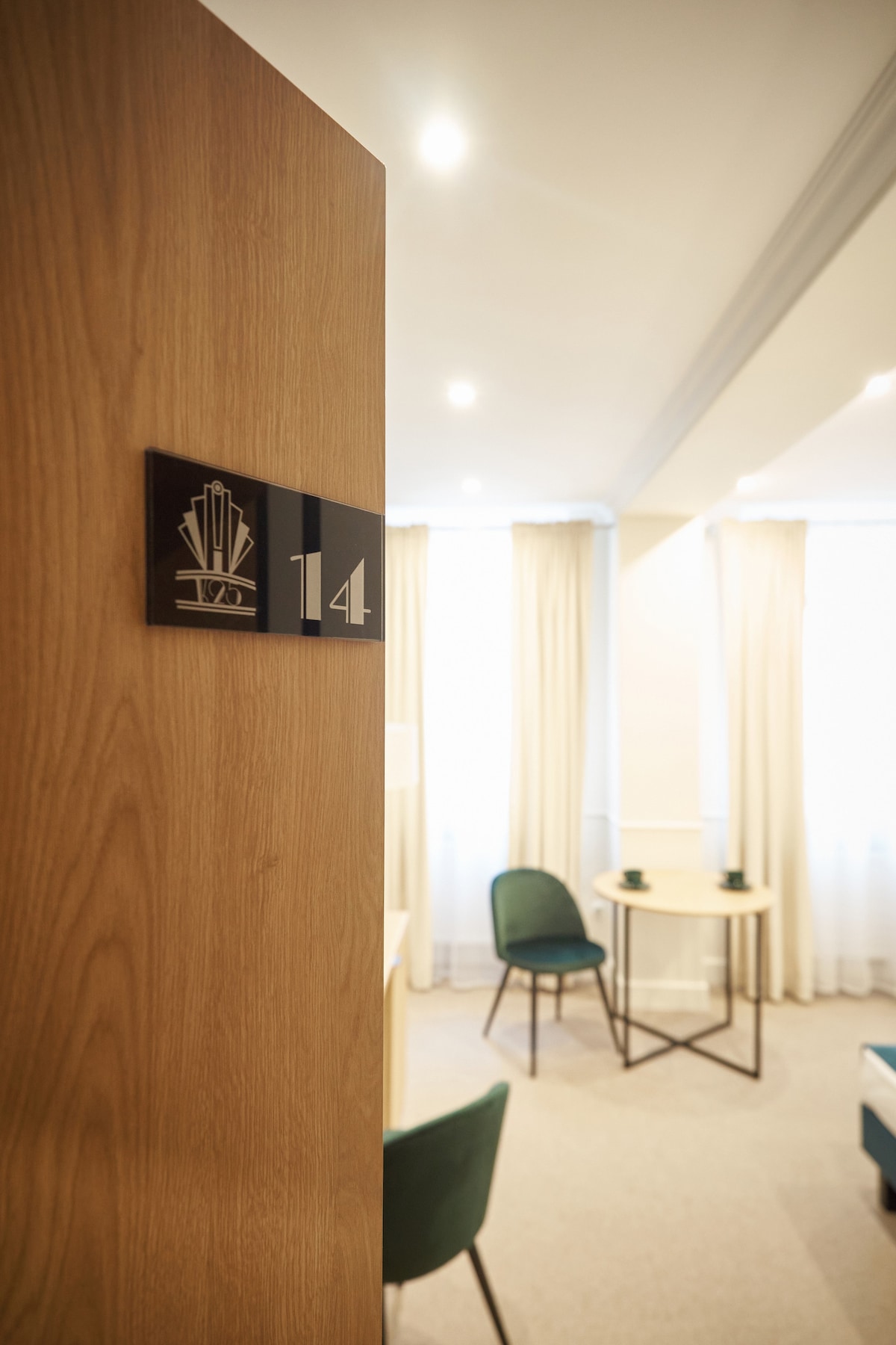 The entrance to Room 14 is framed by a wooden door, displaying a sleek number plaque. Inside, a cozy seating area features a small round table and two upholstered chairs. Soft natural light filters through the sheer curtains, contributing to a relaxing atmosphere.