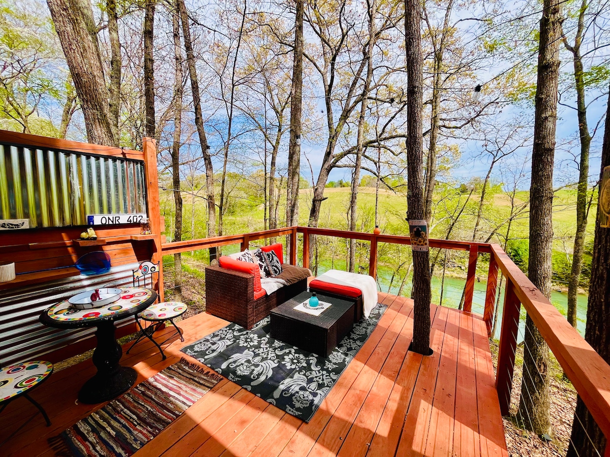 An outdoor deck is nestled among trees, featuring a comfortable seating area with red cushions on dark wicker furniture. A circular table with artistic designs sits nearby, and a textured rug adds warmth. Green hills are visible in the distance, creating a serene backdrop.