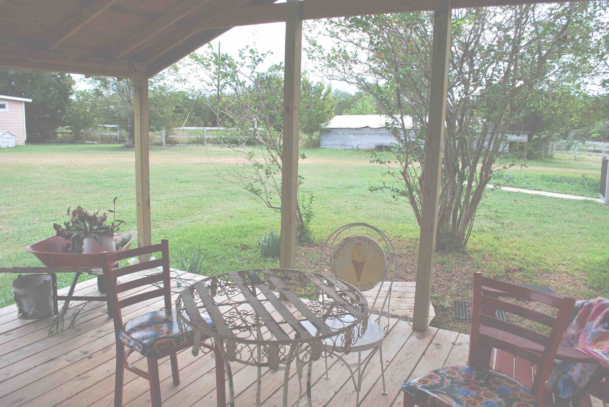 A private porch features a metal table surrounded by four chairs, providing a shaded space for relaxation. The view consists of an expansive grassy area and distant trees, with a small structure and a garden wheelbarrow visible in the background.