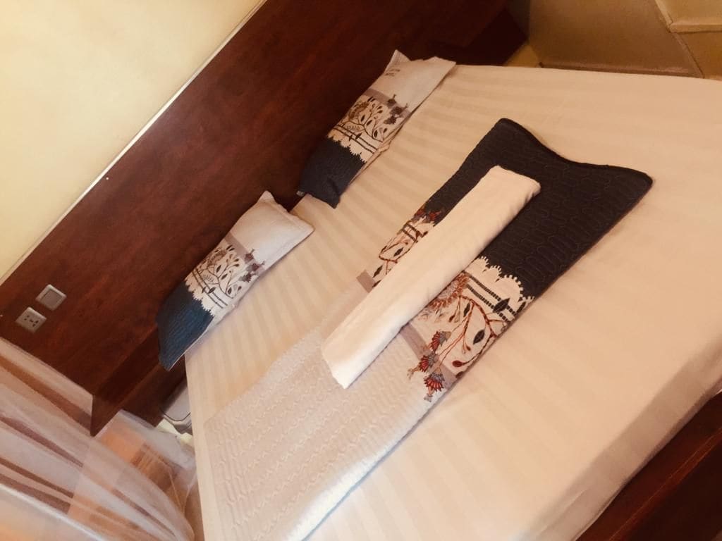 A spacious bed is adorned with decorative pillows and neatly arranged towels. Soft linens cover the bed, complemented by a wooden headboard. The warm-toned walls and subtle lighting create a cozy and inviting ambiance.