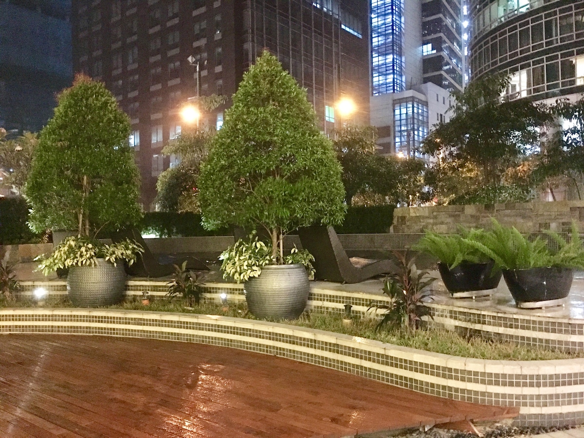 A serene outdoor space features carefully arranged greenery in large planters, with lush plants and neatly trimmed shrubs. The polished wooden deck reflects city lights, enhancing the tranquil setting amidst the surrounding buildings.